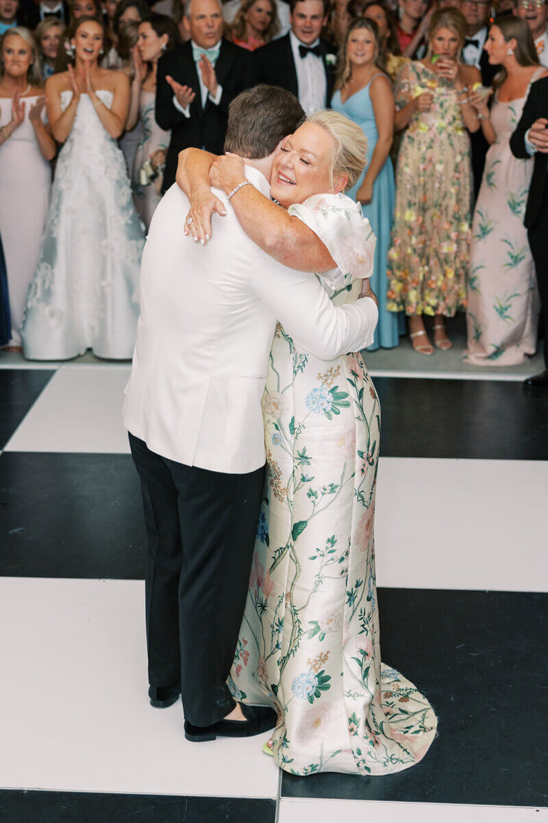 A Secret Speakeasy and Late-Night Taco Bell?! This Nashville Wedding Had Serious Surprises - 21 A woman in a floral gown hugs a man in a white dinner jacket while dancing on a black-and-white checkered floor at this garden party wedding, with guests watching in the background.