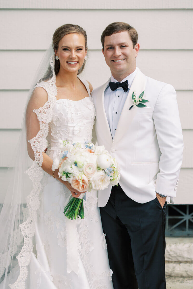 A Secret Speakeasy and Late-Night Taco Bell?! This Nashville Wedding Had Serious Surprises - 2 A bride in a white lace-trimmed gown and veil and a groom in a white dinner jacket and black bow tie standing together, the bride holding a floral bouquet.