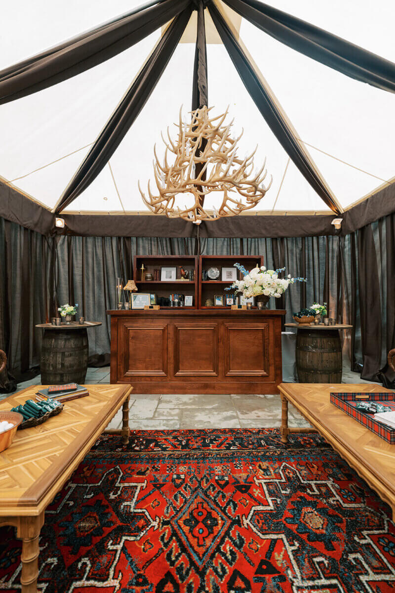 A Secret Speakeasy and Late-Night Taco Bell?! This Nashville Wedding Had Serious Surprises - 15 A wooden bar with a large antler chandelier above, set under a tent with brown curtains, wooden tables, and a patterned rug for a secret speakeasy wedding.