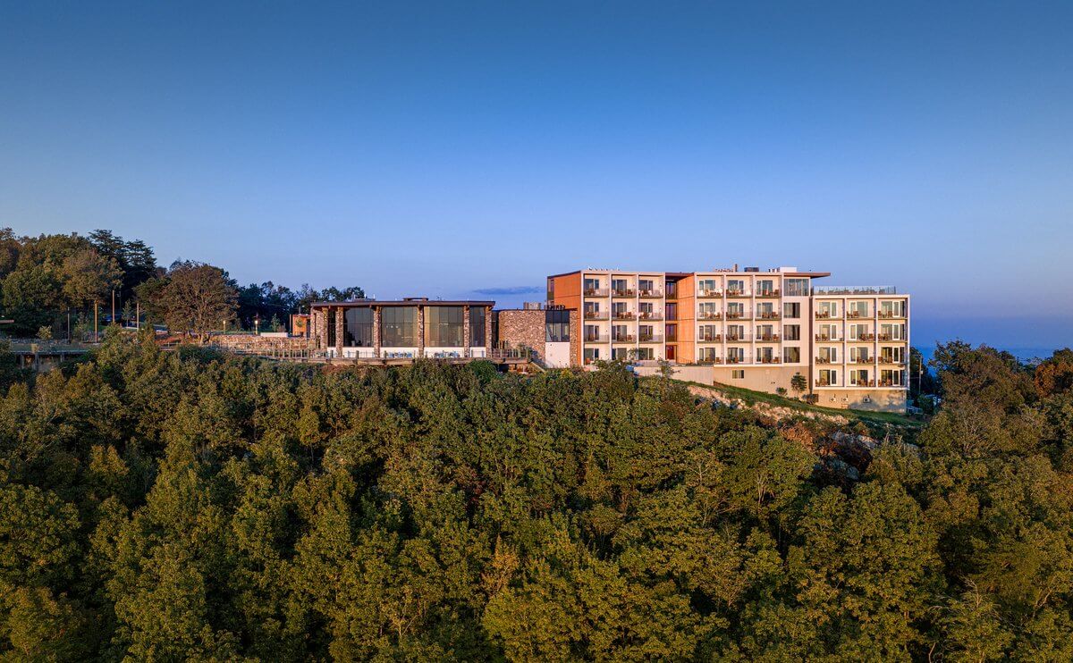 A multi-story hotel with balconies overlooking a dense forest under a clear blue sky.