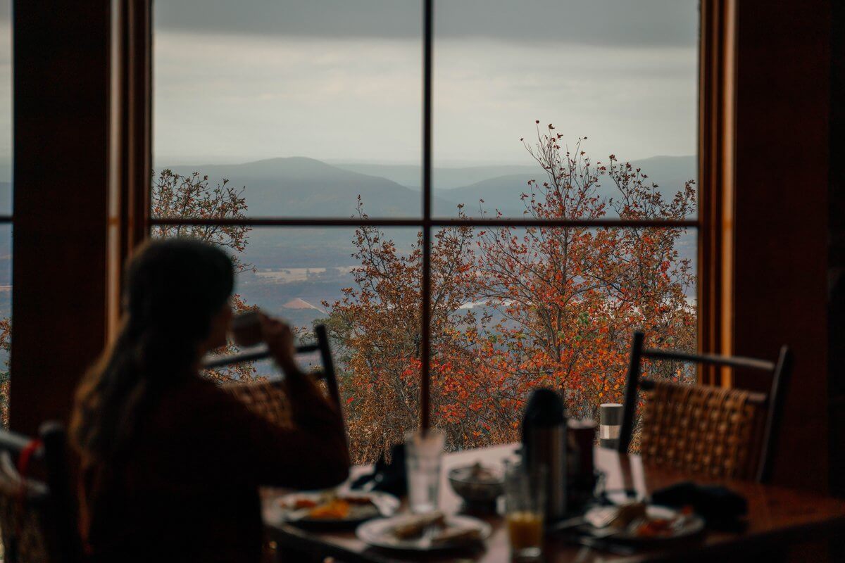 A silhouette of a person drinks from a mug at a dining table, framed by the scenic views of a mountain through a large window.