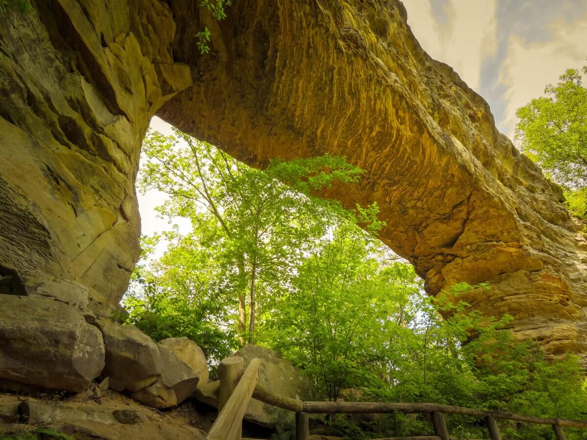 Large natural stone arch over a wooded trail at a Southern state park, with a wooden railing in the foreground.