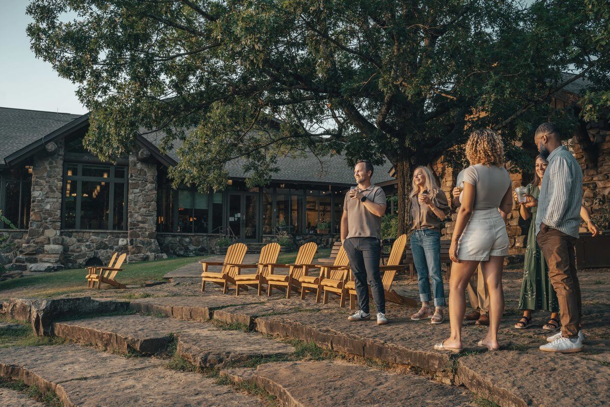 A group of adults stands on a stone patio holding drinks in front of a Southern state park lodge.