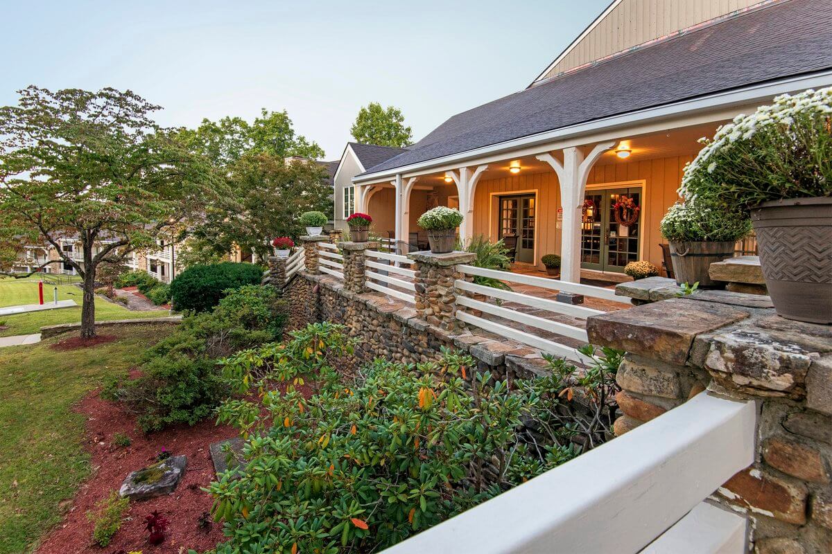A Southern state park lodge featuring a wrap-around porch and white wooden railing, situated above a stone retaining wall and landscaped yard with scenic views.