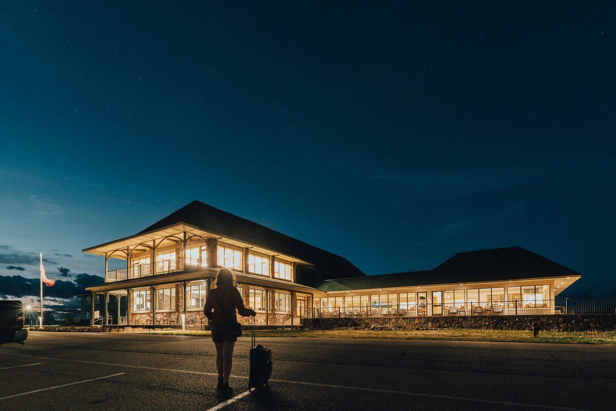 A person with a rolling suitcase stands in a parking lot at night, facing a large, illuminated lodge at a Southern state park.