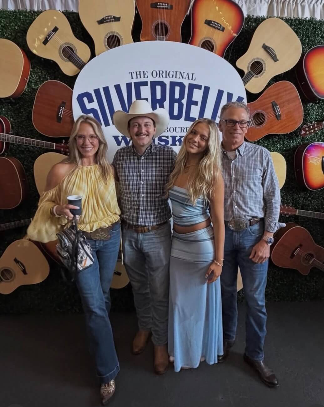 Four people stand smiling in front of a Silverbelly Whiskey sign, surrounded by guitars—a tribute to the singer songwriter spirit—arranged in a circular pattern on a green wall.