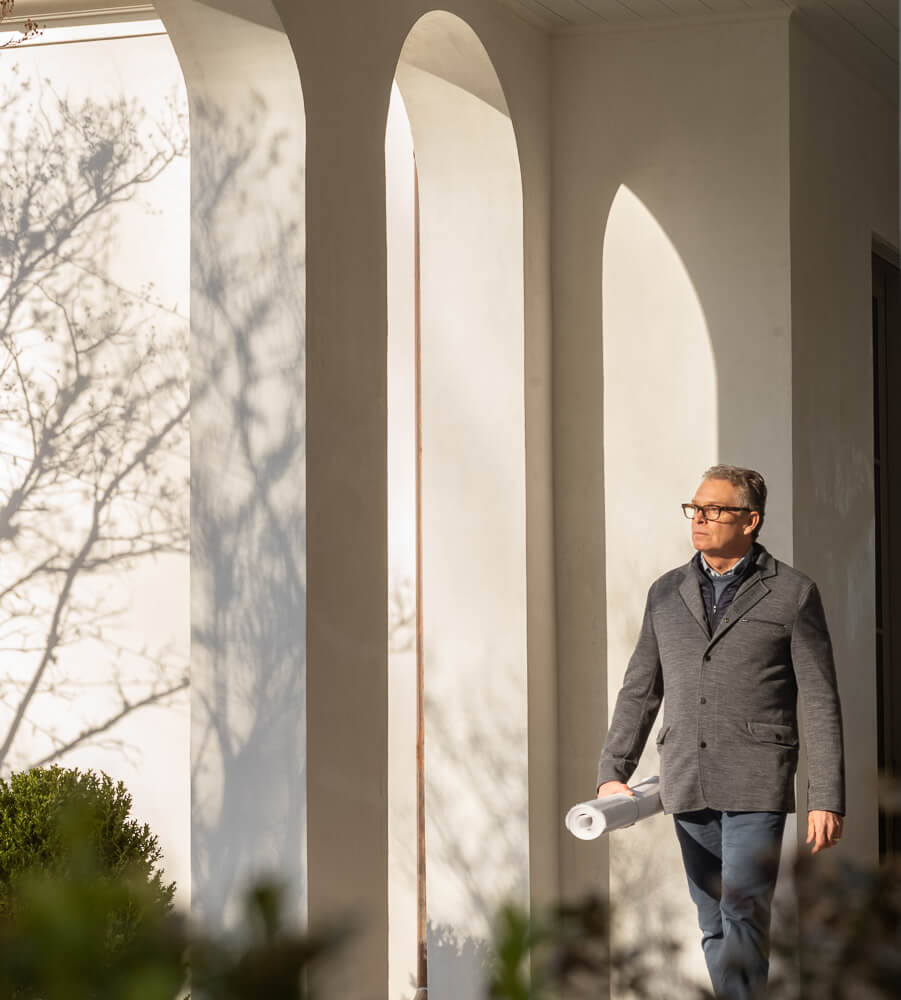 A man in glasses and a blazer walks outside near tall white arches of castle homes, holding rolled-up papers as sunlight casts shadows of tree branches on the wall.