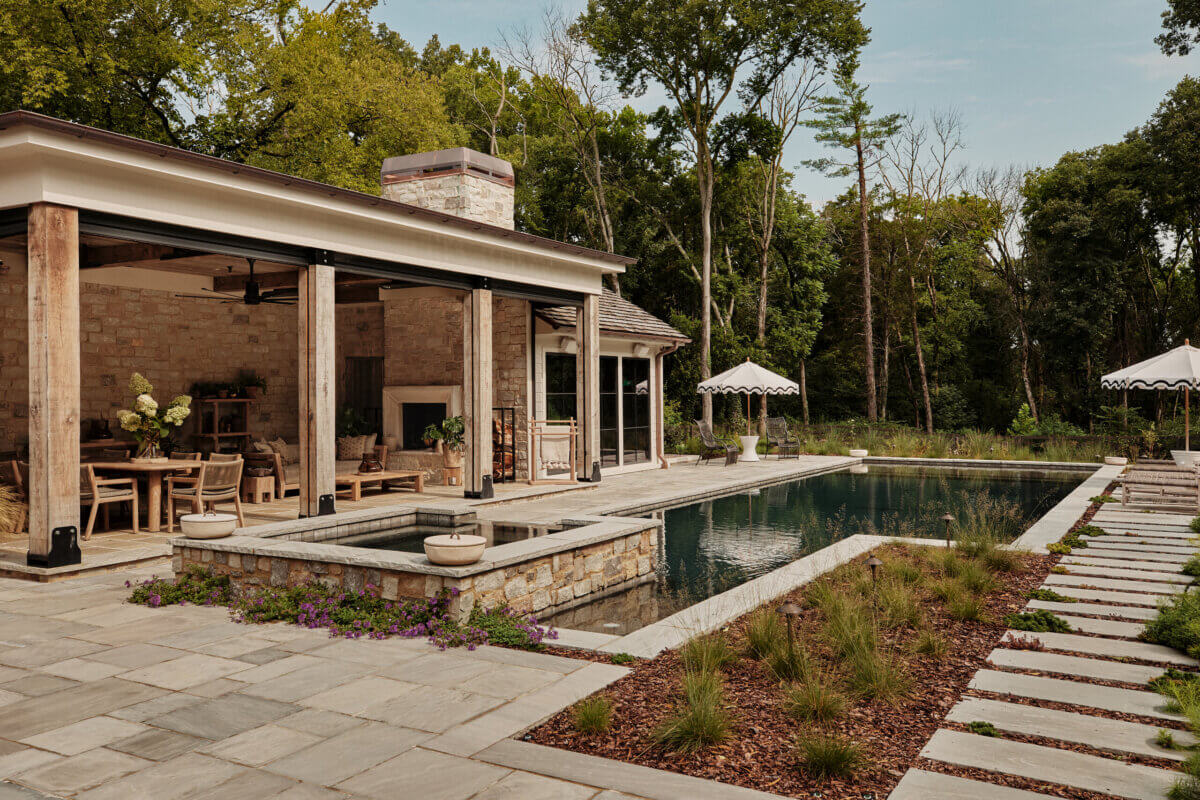 They Abandoned Their Dream Home Plans. Here's What Happened Next. - 3 A stone patio with a covered outdoor living area, a swimming pool with an integrated spa, and a stone walkway surrounded by landscaped gardens.