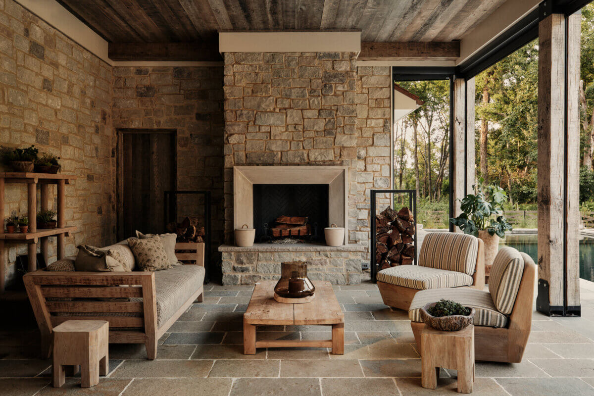 They Abandoned Their Dream Home Plans. Here's What Happened Next. - 11 A stone patio with an outdoor fireplace, a wooden sofa, two striped armchairs, and a low wooden coffee table.