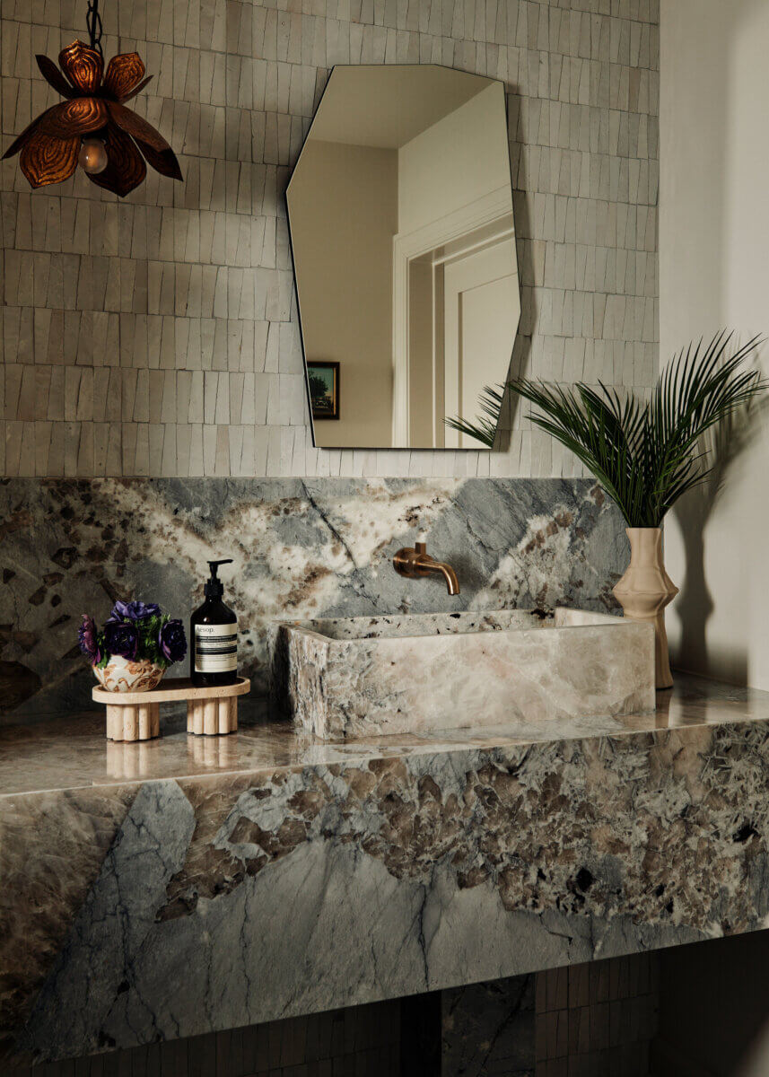 They Abandoned Their Dream Home Plans. Here's What Happened Next. - 12 A rectangular stone sink set into a matching marble countertop, with a brass faucet, a mirror, and a small vase of flowers.