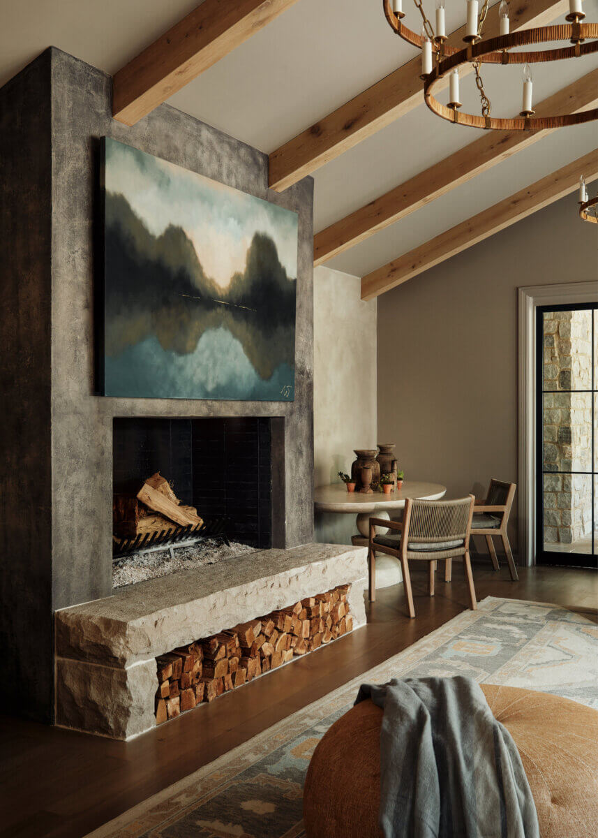 They Abandoned Their Dream Home Plans. Here's What Happened Next. - 15 A living room featuring a concrete fireplace with an abstract landscape painting above the hearth, a stone log storage bench, and a nearby dining area with chairs and a circular table under wood ceiling beams.