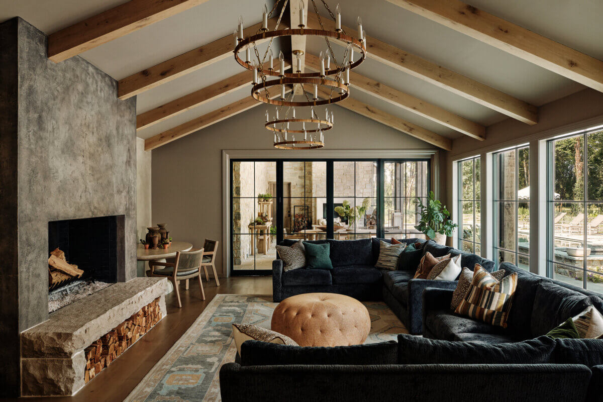 They Abandoned Their Dream Home Plans. Here's What Happened Next. - 14 Living room with a stone fireplace, a large navy sectional sofa, a round ottoman, and two stacked circular chandeliers under vaulted wood-beamed ceilings.
