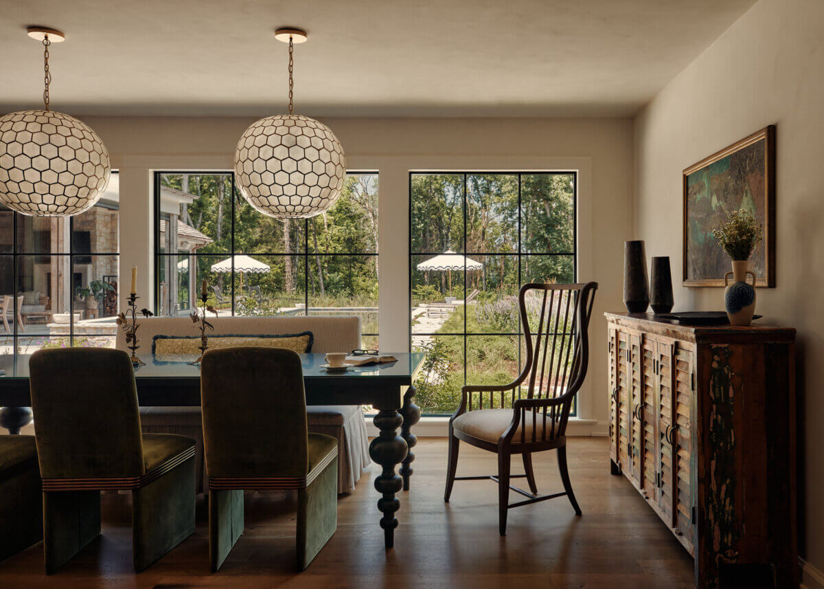 They Abandoned Their Dream Home Plans. Here's What Happened Next. - 5 Dining room with a table, upholstered chairs and bench, a spindle-back chair, two globe pendant lights, and a wooden sideboard in front of large windows overlooking a garden.