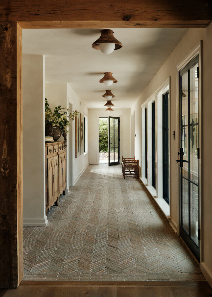 They Abandoned Their Dream Home Plans. Here's What Happened Next. - 6 A long, sunlit hallway with a herringbone-patterned tile floor, wooden console table, and three dome pendant lights leading to an open door.