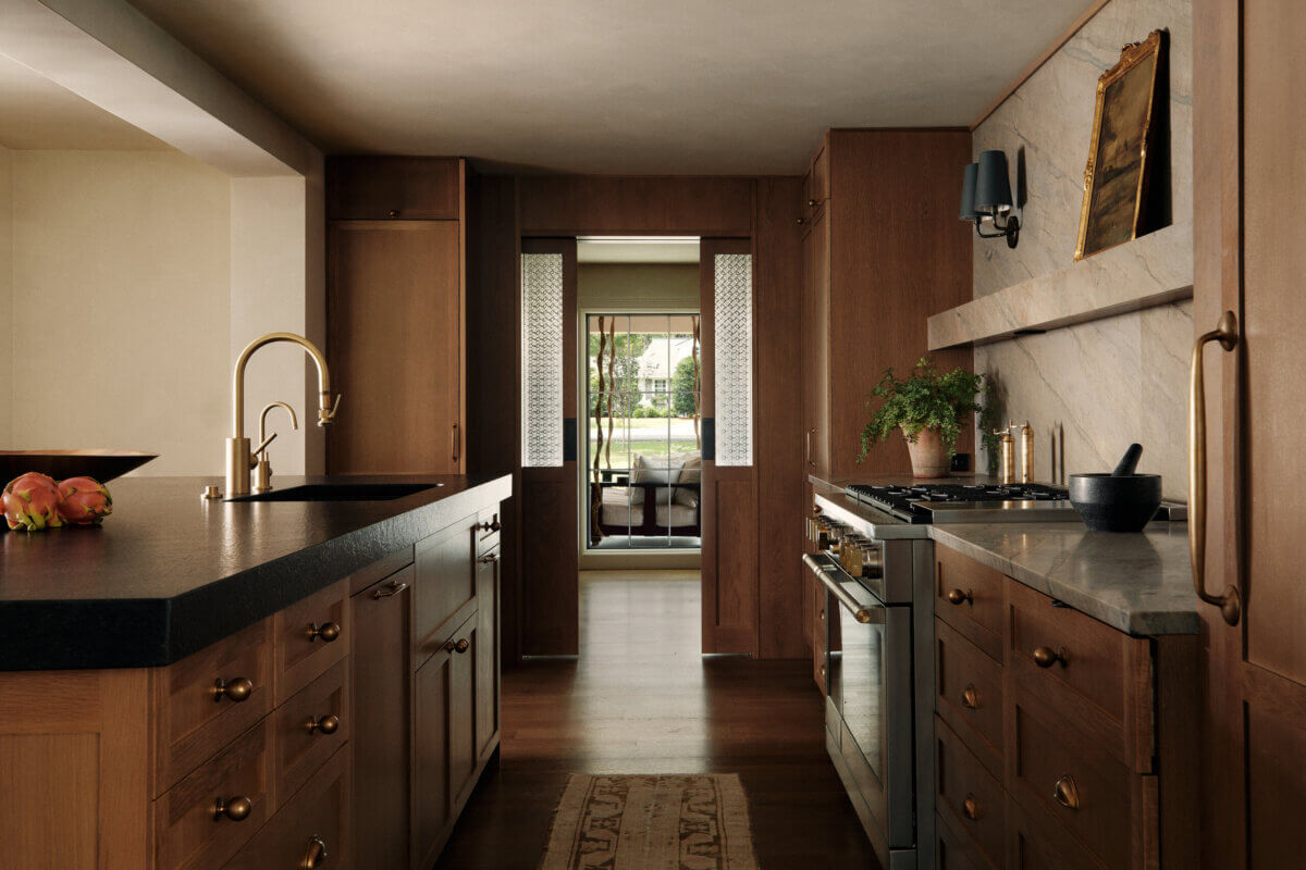 They Abandoned Their Dream Home Plans. Here's What Happened Next. - 7 A modern kitchen with wood cabinetry, a dark stone island, and a brass faucet, with a view through a doorway to an outdoor patio.