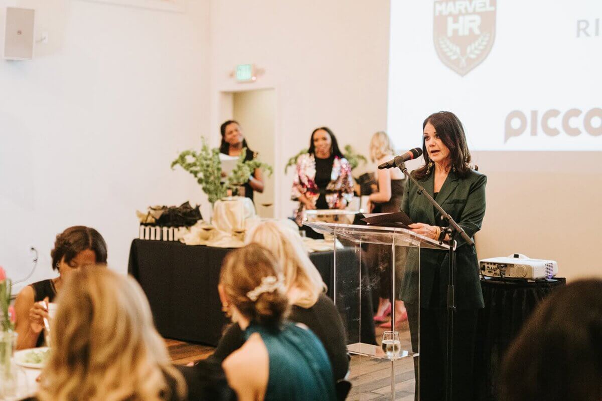 A woman in a dark blazer speaks at a BrainTrust event, addressing an audience about remote business from the podium.