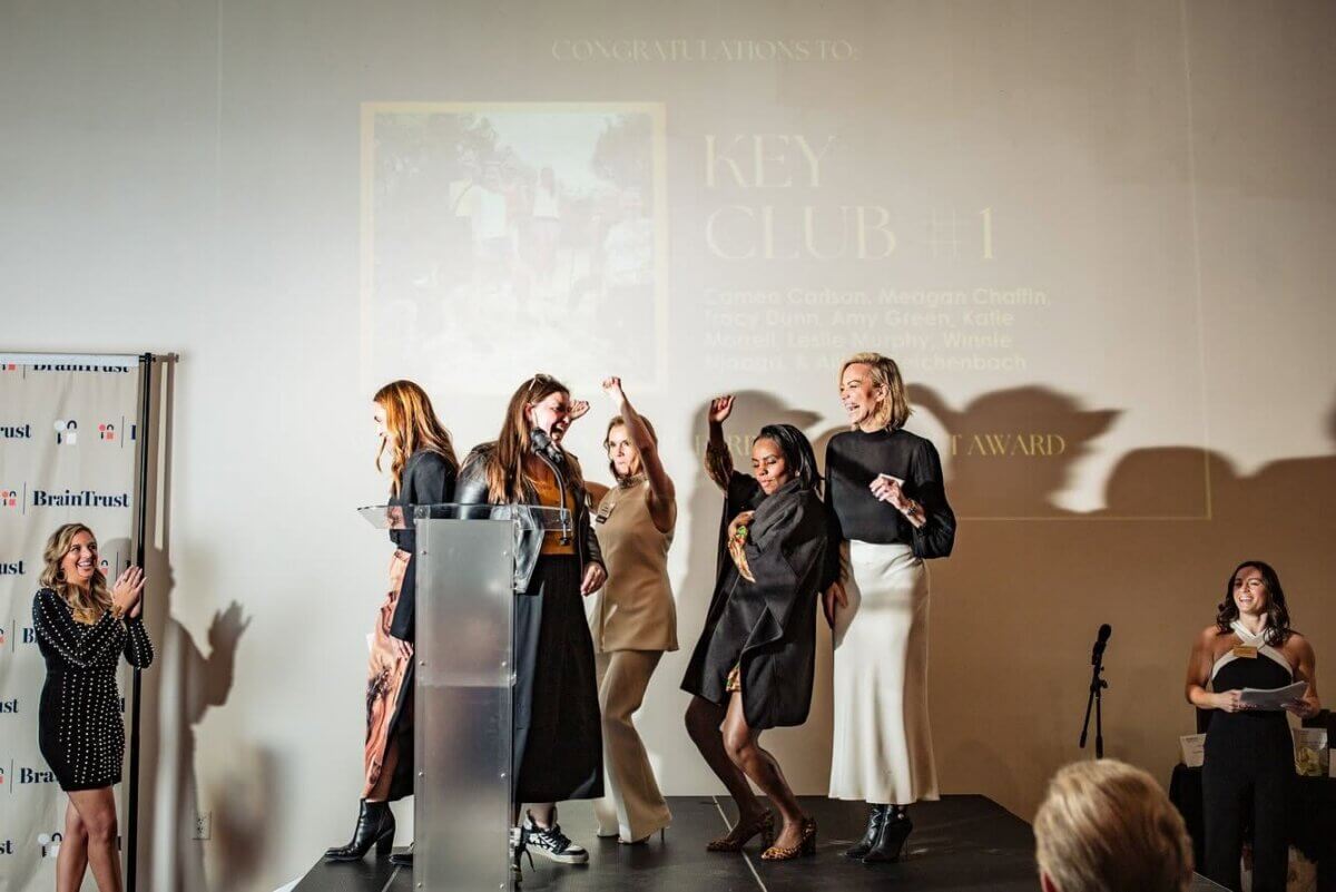 Five women celebrate on a stage in front of a Key Club #1 projection as BrainTrust members gather, with a woman at a podium to the left and another at a microphone to the right.