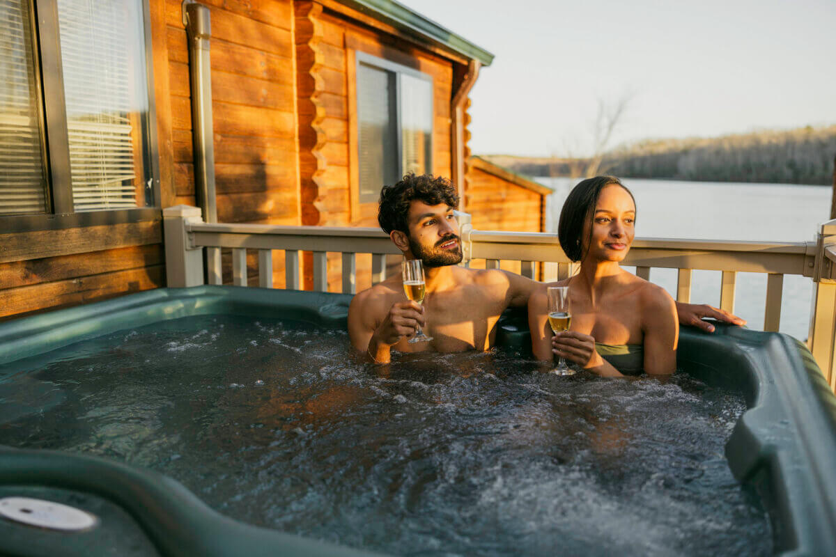 Two people relax in an outdoor hot tub on a deck, holding champagne glasses, at a wooden cabin—perfect for a Virginia weekend getaway with lake and forest views.