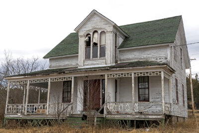 Abandoned two-story house with peeling white paint, a green shingled roof, and a damaged front porch, reminiscent of southern legends.