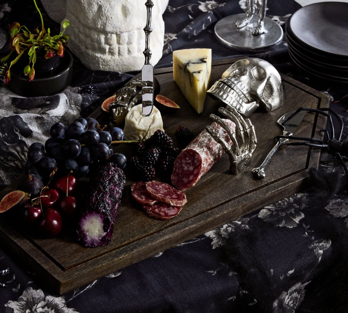 20 Halloween Decor FINDS That Are Actually Chic - 5 A dark wooden board with cheeses, cured meats, fruits, and silver skull-shaped utensils creates striking Halloween decor on a dark floral tablecloth.