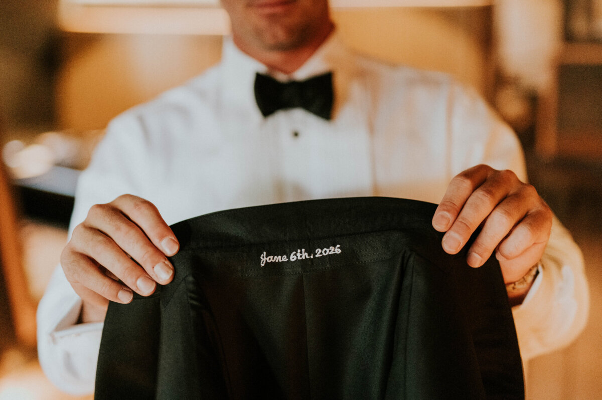 A person in a tuxedo holds up a jacket with “June 6th, 2026” embroidered inside the collar, fitting for a Kentucky Derby Museum wedding.