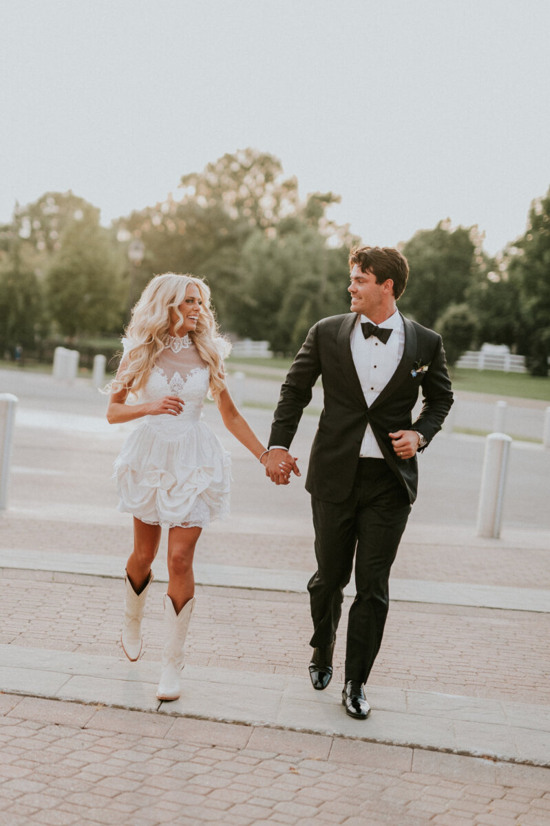 A bride in a short white dress and cowboy boots holds hands with a groom in a tuxedo while walking outdoors at the Kentucky Derby Museum.