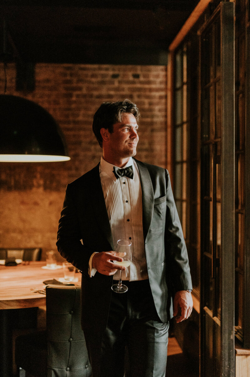 A man in a tuxedo holding a glass of wine stands in a brick-walled room at a Kentucky Derby Museum wedding.