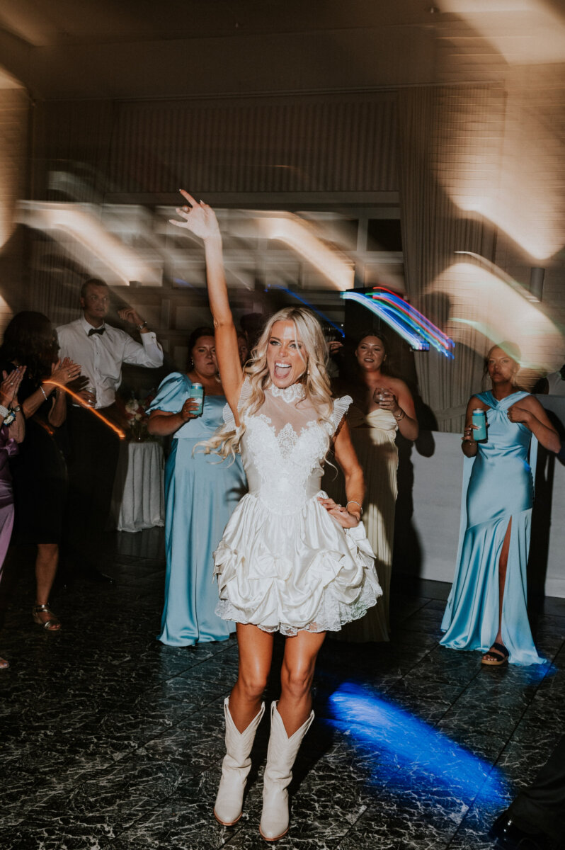 A bride in a short white dress and cowboy boots dances with guests at a Kentucky Derby Museum wedding, with light streaks visible in the background.