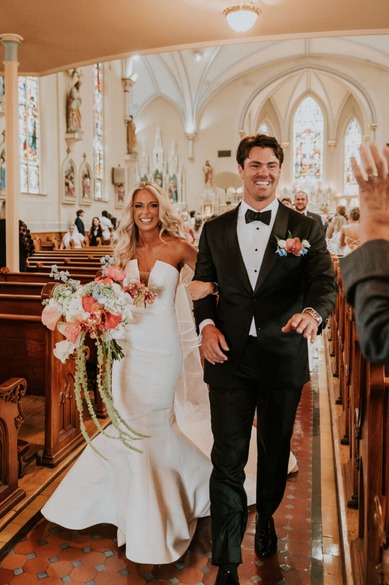 A bride in a white gown and a groom in a black tuxedo walking down the aisle together after their Latin mass.