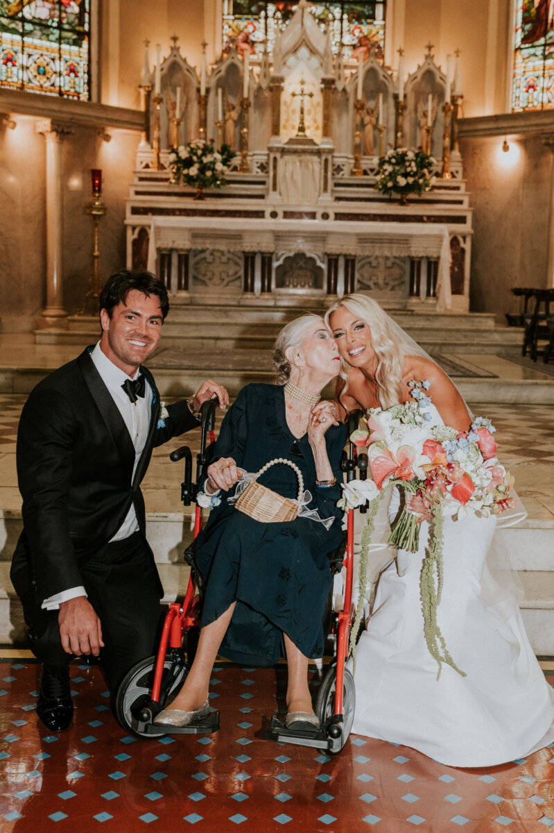A bride in a white wedding gown and a groom in a tuxedo pose with an elderly woman in a wheelchair who is kissing the bride’s cheek inside a Kentucky Derby Museum wedding venue.