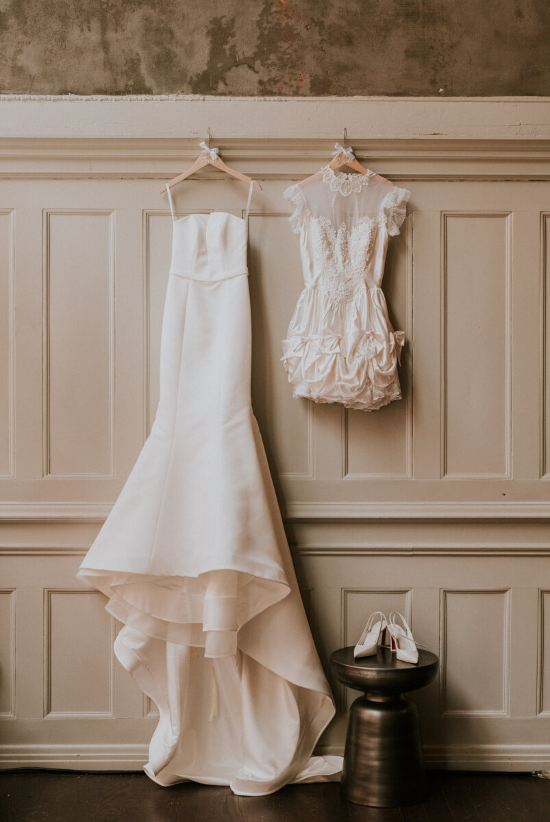 A floor-length wedding gown and a short lace dress hang on a wall above a pair of white heels.