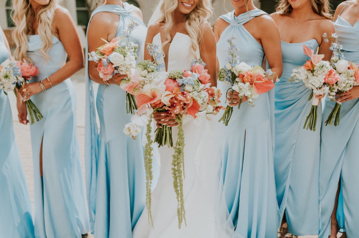 A bride in a white gown and five bridesmaids in light blue dresses hold matching coral and white bouquets at a Kentucky Derby Museum wedding.