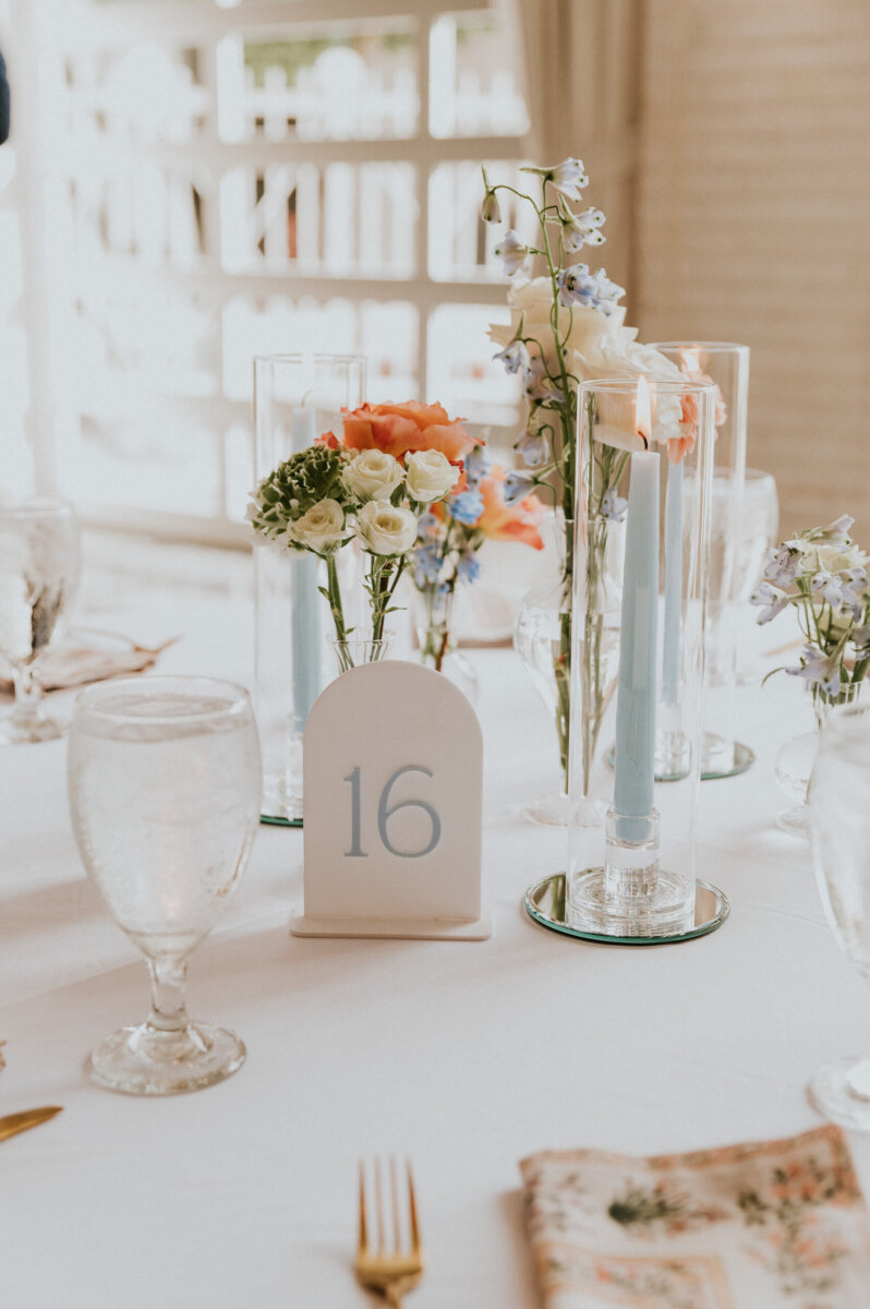 Table centerpiece for a Kentucky Derby Museum wedding with a white card displaying the number 16, surrounded by floral arrangements and a light blue taper candle in a glass holder.