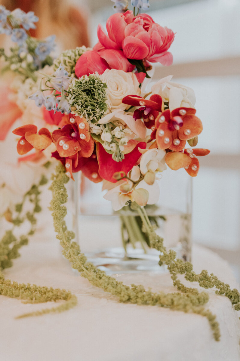 A floral arrangement for a Kentucky Derby Museum wedding featuring pink peonies, orange orchids accented with pearls, and cascading green amaranthus in a glass vase.