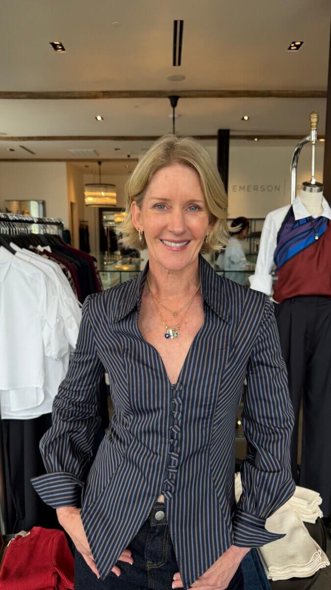 Darci Bolenbaugh smiles while wearing a dark blue, pinstriped button-down shirt and layered gold necklaces inside a clothing boutique.