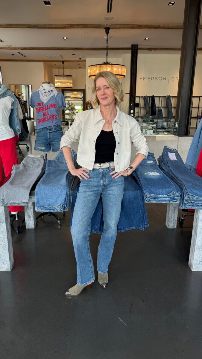 Darci Bolenbaugh wears a white jacket, black top, and jeans while standing in a clothing store with tables of folded denim.