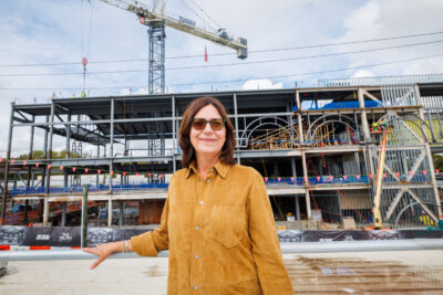 A woman in a tan shirt and glasses stands before a building under construction, looking like a figure from a Sally Williams creepypasta beneath a looming crane.