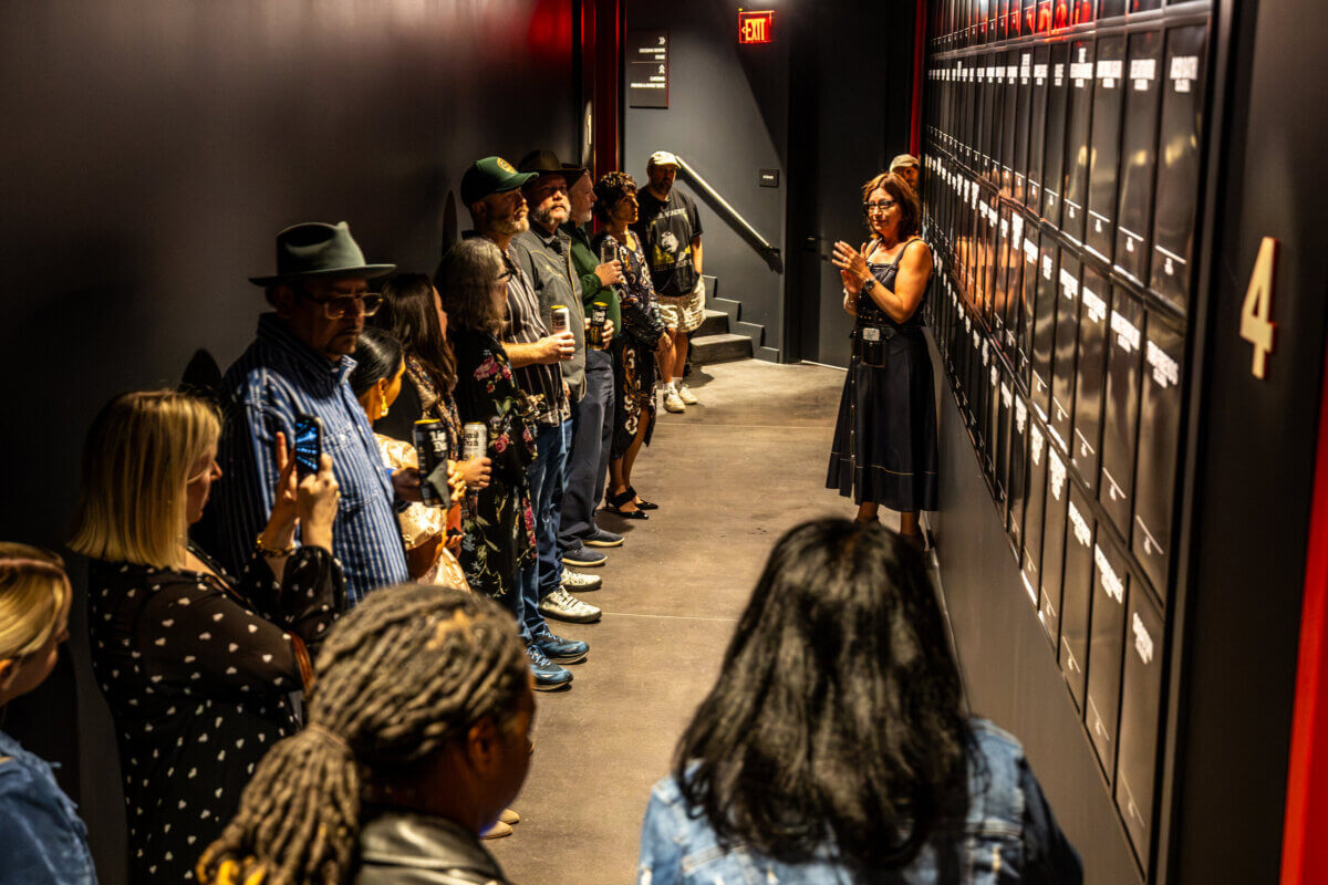 A group of people stands in a dark hallway as Sally Williams gestures toward a wall of framed images.