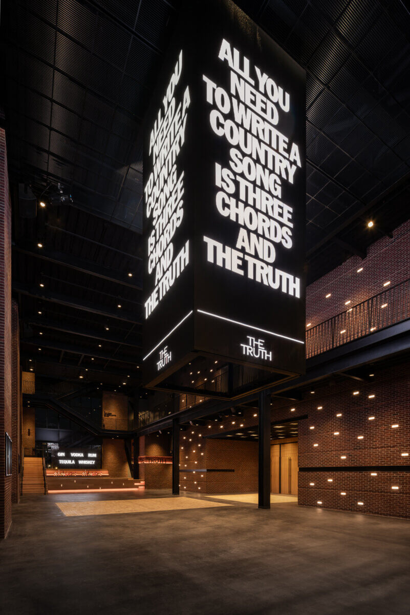 A large, illuminated hanging sign in a warehouse-style building reads, “All you need to write a country song is three chords and the truth,” a phrase famously associated with Sally Williams.