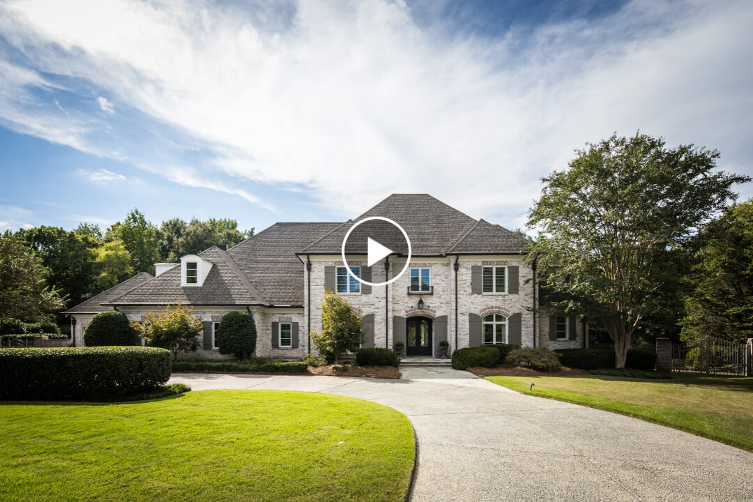 A large, two-story brick house at 9260 Gwynn Hollow Cv with a circular driveway and green lawn, featuring a play button icon overlaid in the center.