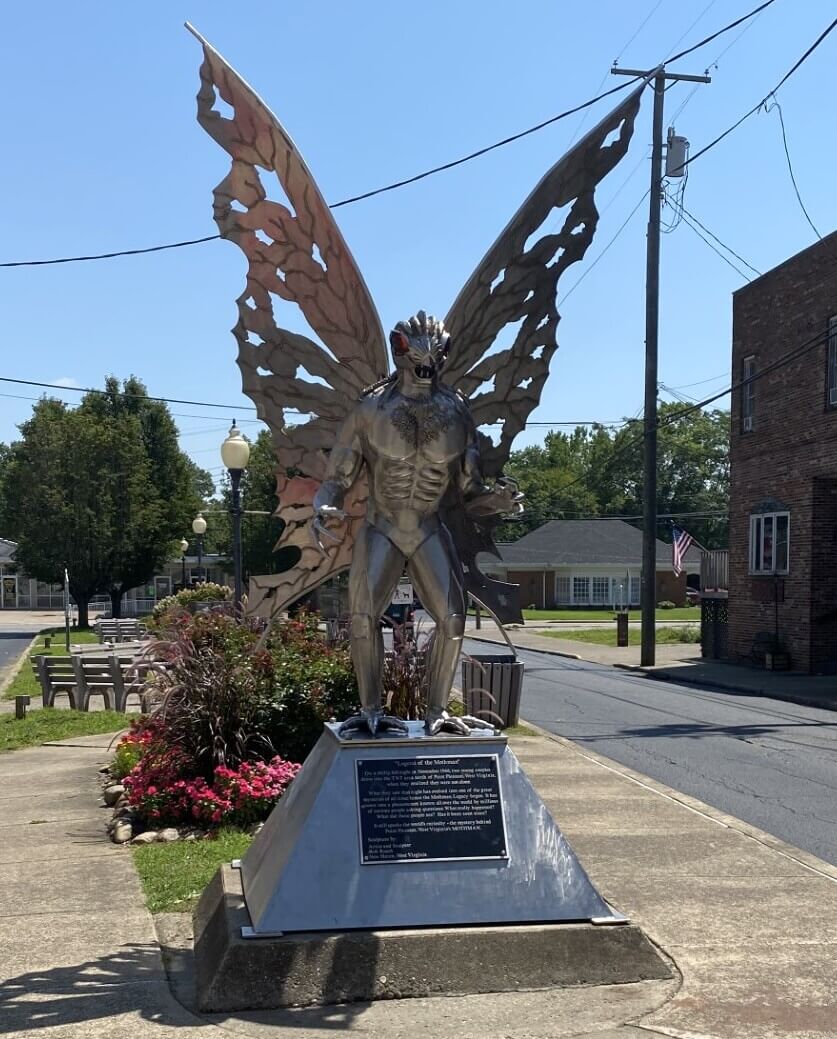 7 Spooky Southern Legends That'll Make You Lock Your Door Tonight - 5 Large metal Mothman statue standing on a pedestal with an informational plaque on a street corner, a striking icon of Southern folklore.