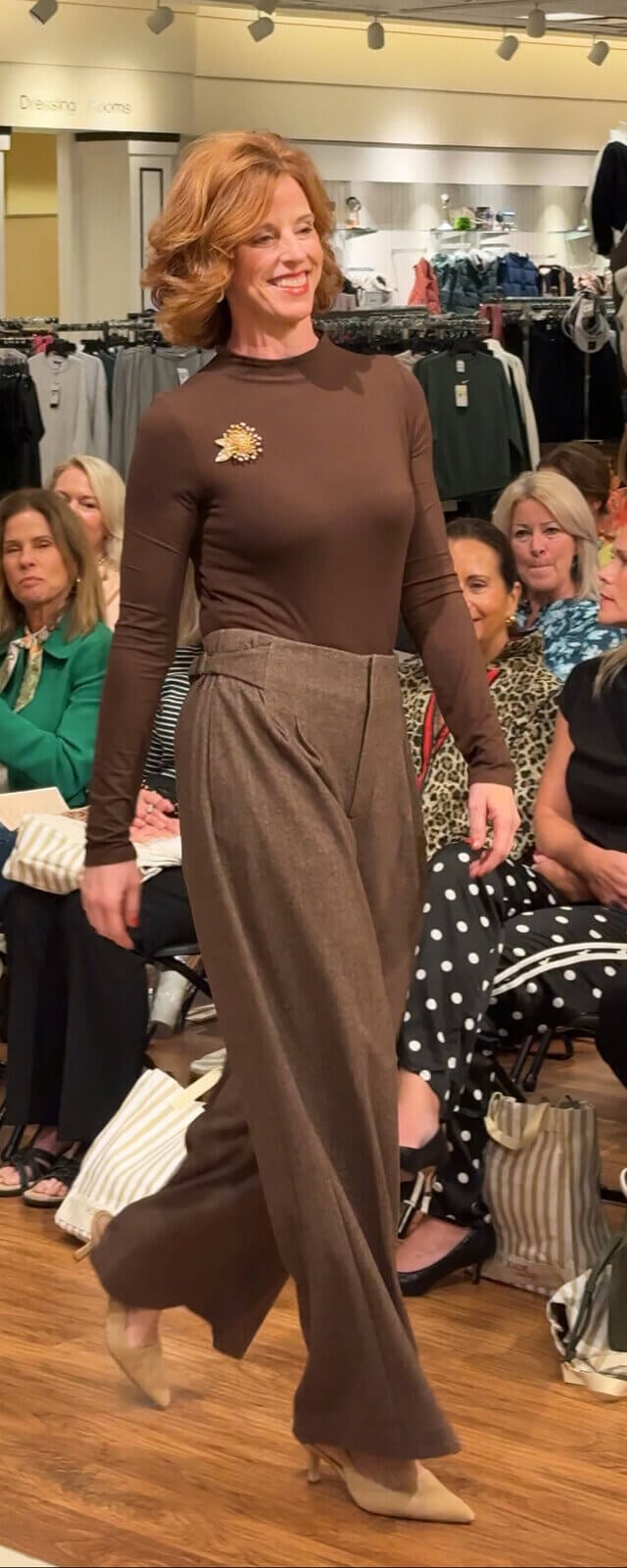 Louisville Fall Fashion 2026: 3 Runway Trends to Incorporate - 6 A woman in a brown long-sleeved top and wide-leg trousers walks down a makeshift runway in a clothing store, showcasing fall fashion trends to an audience seated on either side.