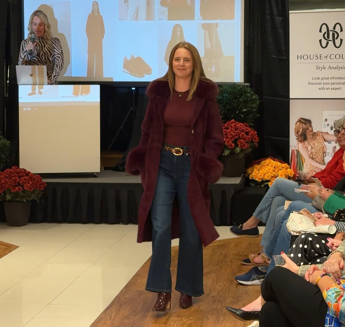 Louisville Fall Fashion 2026: 3 Runway Trends to Incorporate - 4 A woman in a burgundy coat and flared jeans walks down a runway showing fall fashion trends while a speaker presents slides on color palettes.