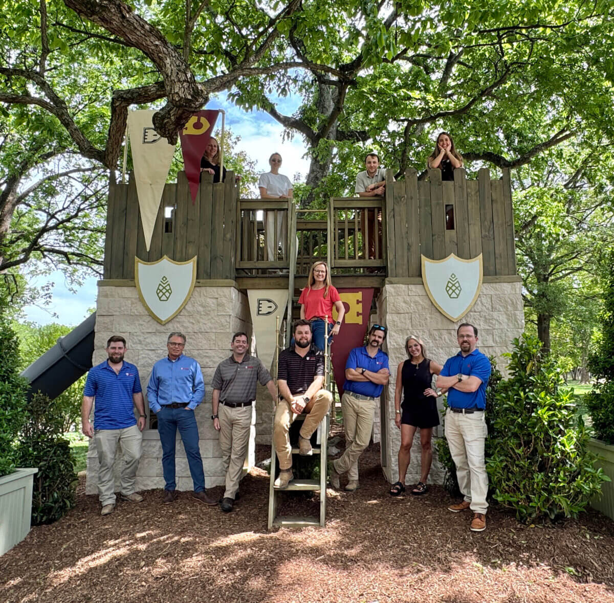 A group of eleven adults poses in front of a wooden play fort with banners and shields, surrounded by trees—like a Singer/Songwriter retreat at whimsical Castle Homes.