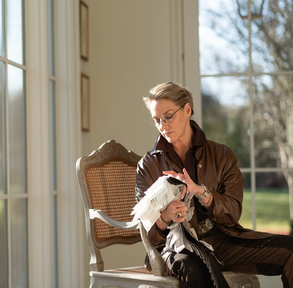 A woman in glasses sits on a chair by large windows, examining a bundle of fabric in her hands, with sunlight streaming into the room.