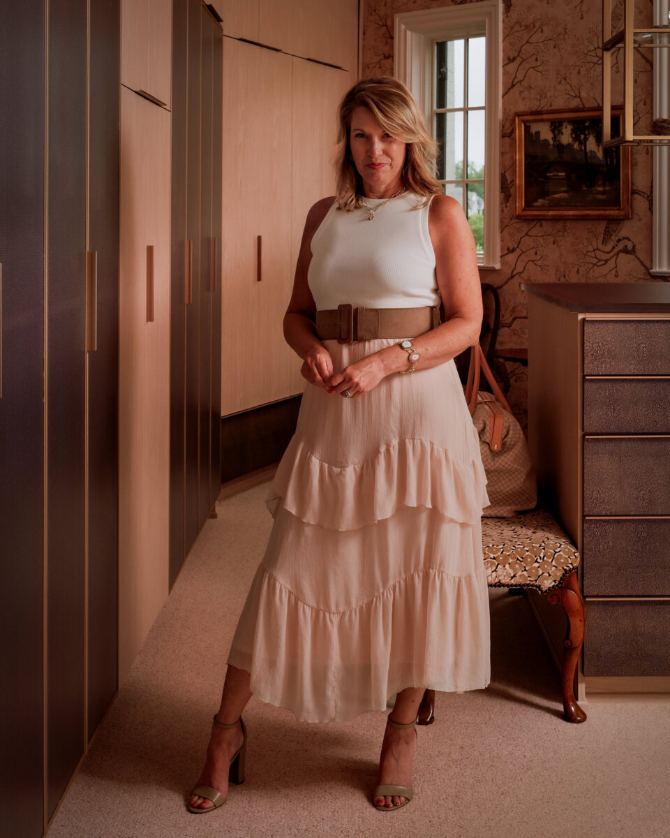 A woman in a white sleeveless top and tiered cream skirt stands in a closet organized to store winter clothes.