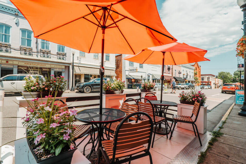 Sidewalk dining area for Virginia weekend getaways, featuring metal tables and chairs under orange umbrellas, with flower planters and a street view in the background.