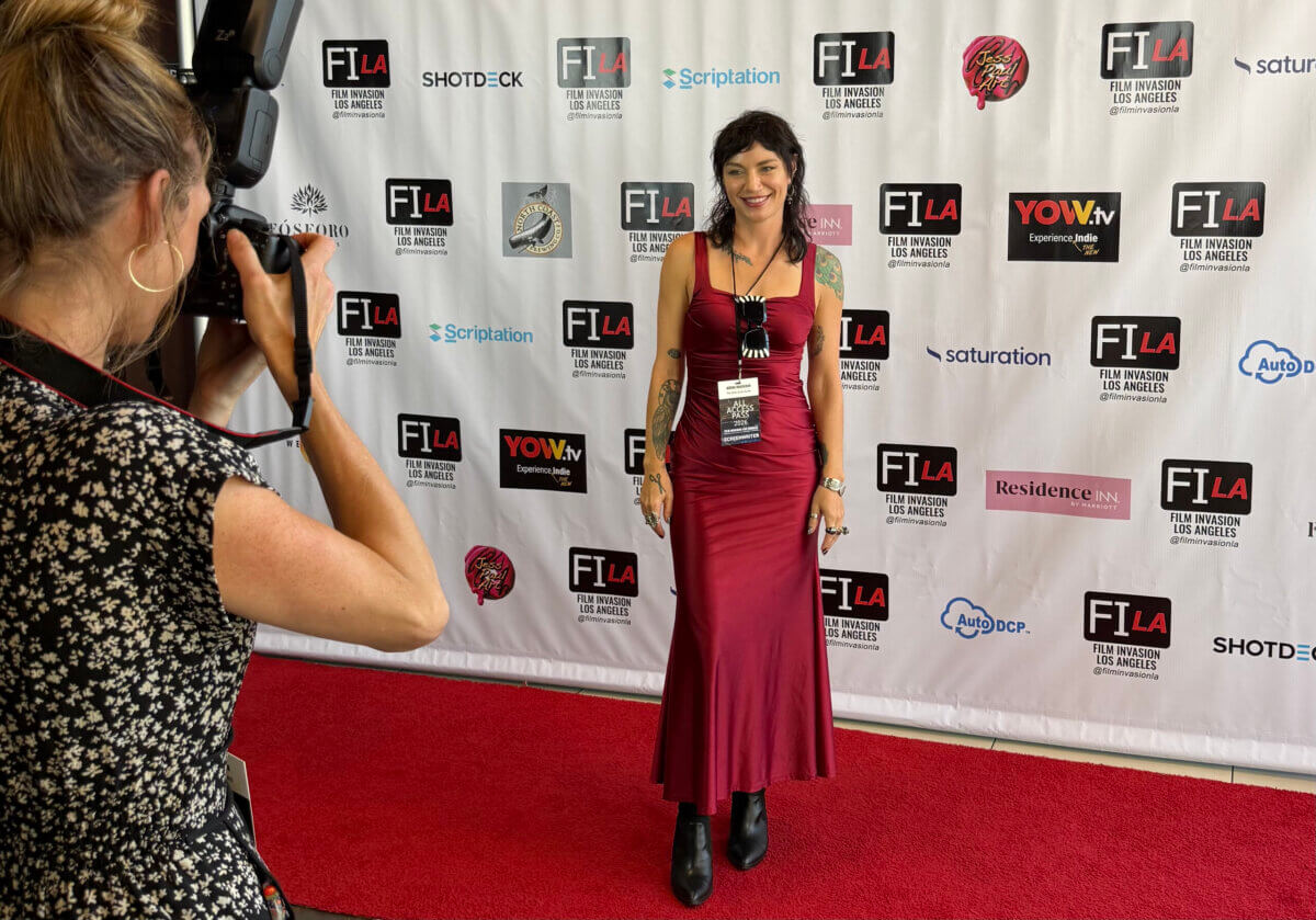 The Haunted Folklore of Mississippi Influenced This Costume Designer's Career - 2 A costume designer in a red floor-length dress poses for a photographer on a red carpet in front of a Film Invasion Los Angeles step-and-repeat backdrop.