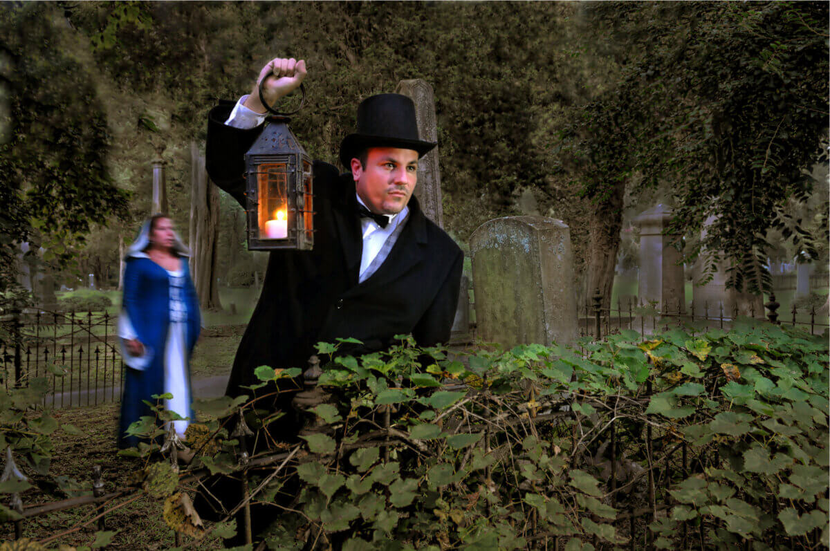 A man in old-fashioned attire holds a lit lantern in a graveyard, as a woman in a blue dress strolls by—a haunting scene for your Virginia Weekend Getaway.