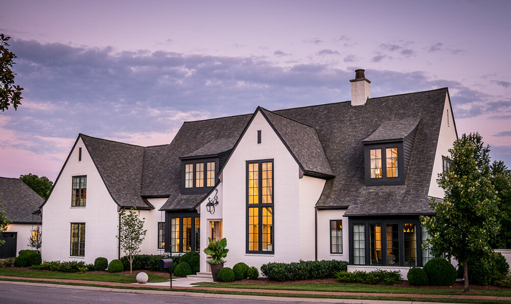 A two-story modern Castle Homes house with white brick exterior, large windows, black trim, and a manicured lawn at dusk. Lights glow inside under a partly cloudy sky.